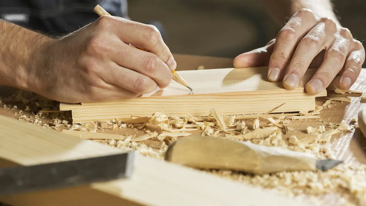 A person using a pencil to mark a wooden piece on a workbench with shavings around.