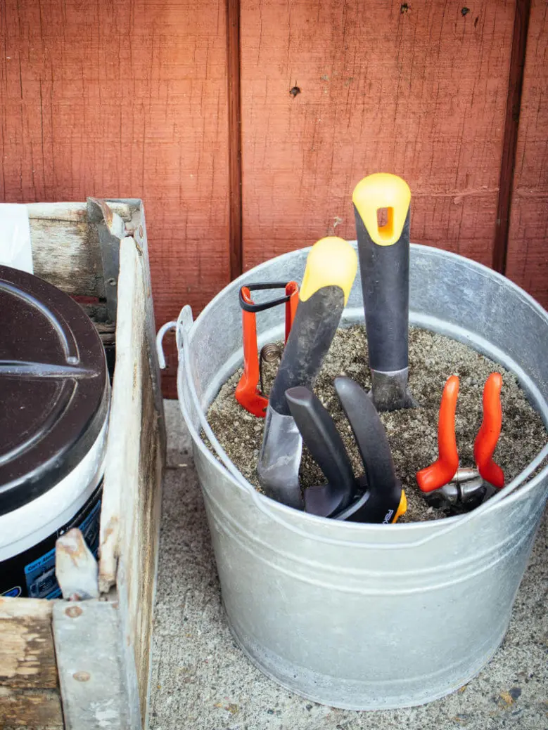 Garden tools embedded in sand within a metal bucket, alongside a wooden crate.