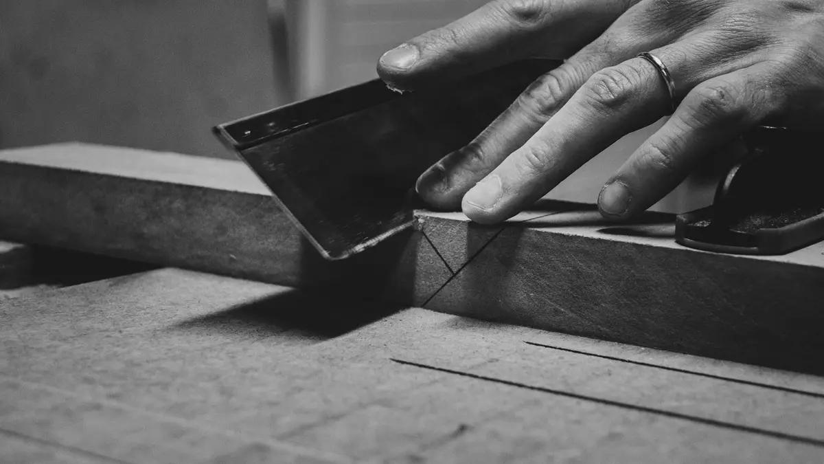 A hand using a metal scraper on a wooden piece, outlining a measurement.